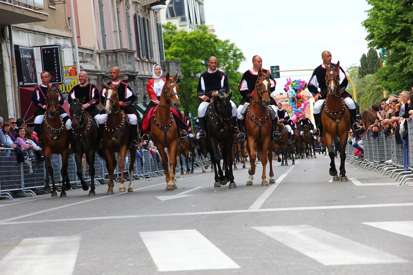Cavalieri in costume tradizionale sardo, con gilet neri bordati di rosso e una donna con velo bianco, sfilano a cavallo lungo un viale di Sassari fra le transenne