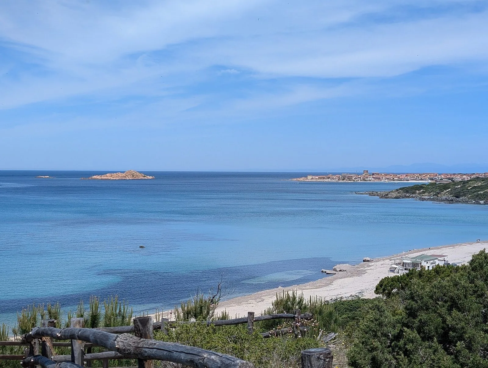 Il borgo dell'Isola Rossa con la torre sul promontorio e l'isolotto rossastro nel mare azzurro, visti dalla costa con una spiaggia chiara in primo piano